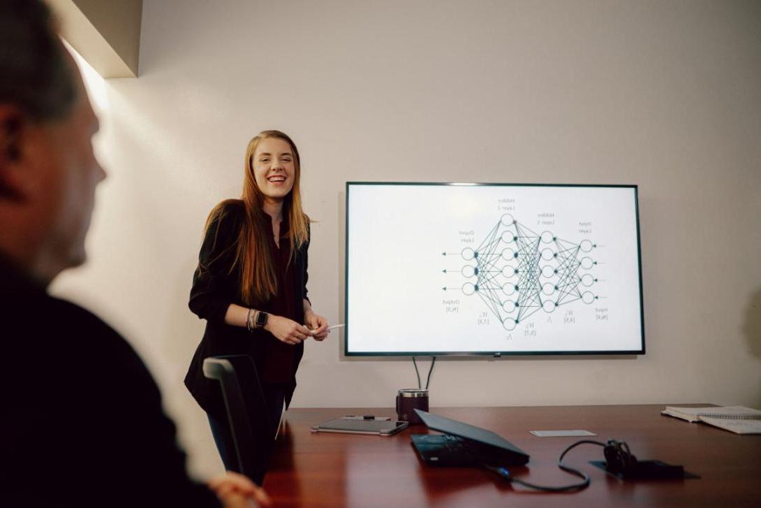 A Kettering School of Management student stands in a conference room. Her presentation is shown via a large monitor.